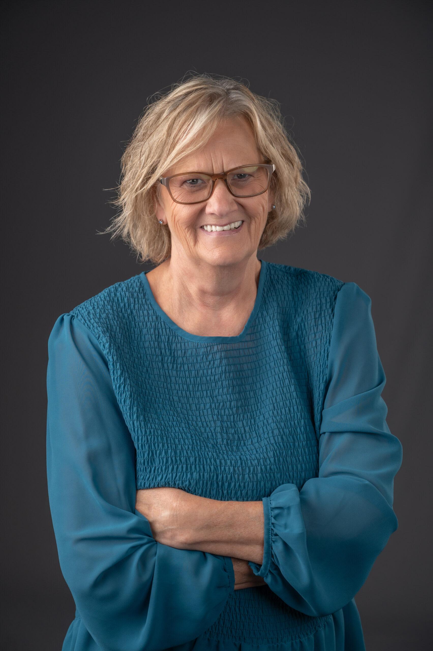 Sue Johns Brooklyn Kenney portrait with grey background while wearing blue top and smiling.