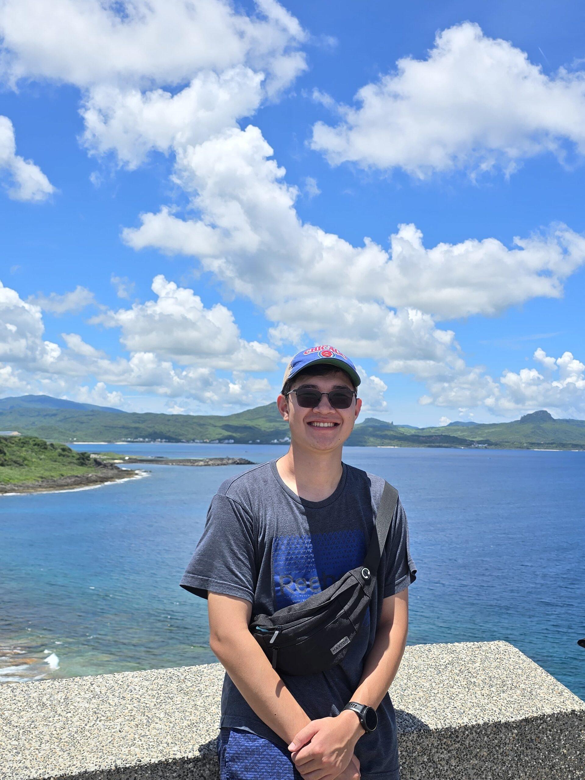A young man wearing a baseball cap and sunglasses stands in front of a large body of water and smiles at the camera.