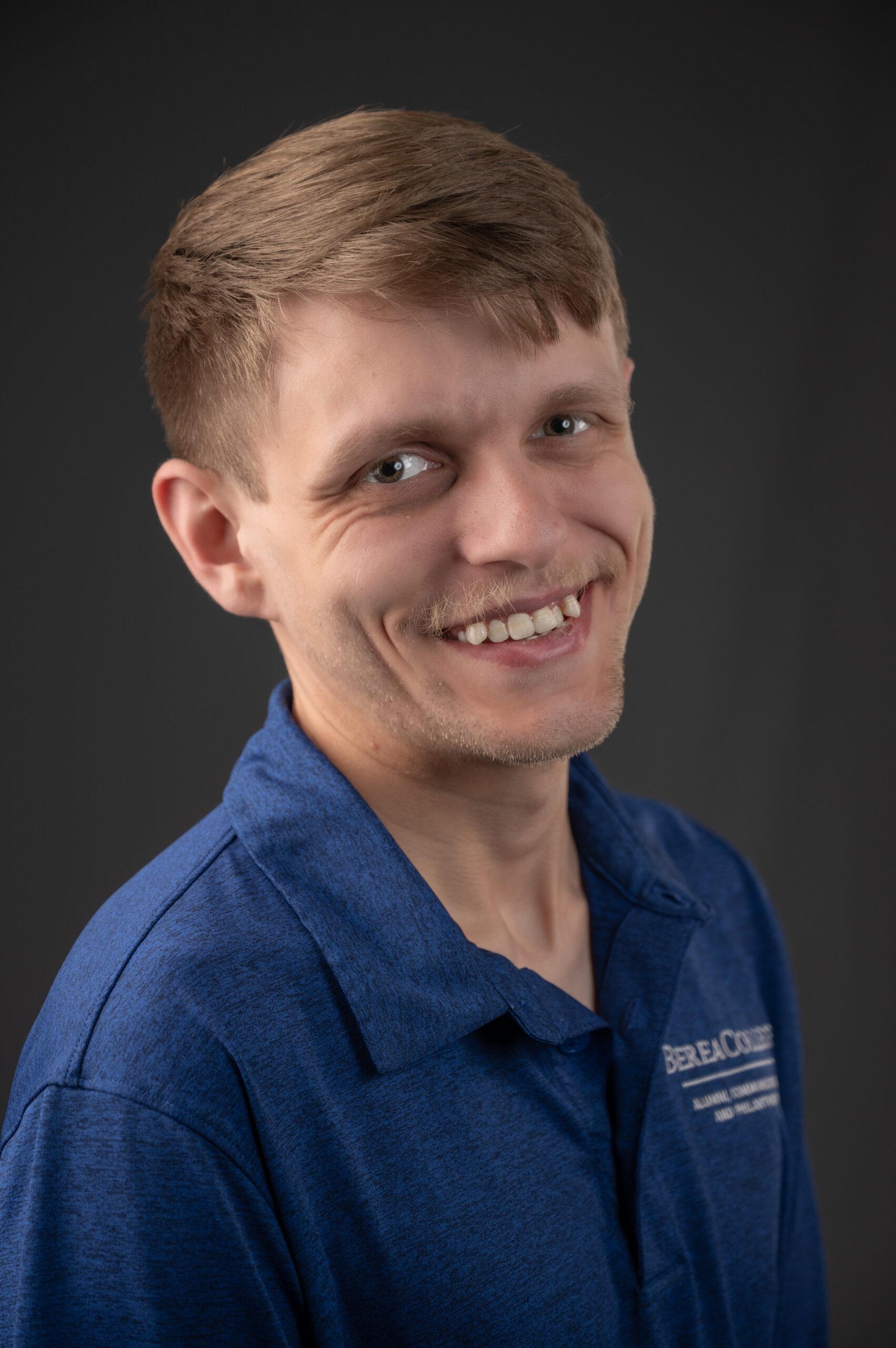 Dustin Cooper headshot smiling towards the camera wearing a blue top and a grey background.
