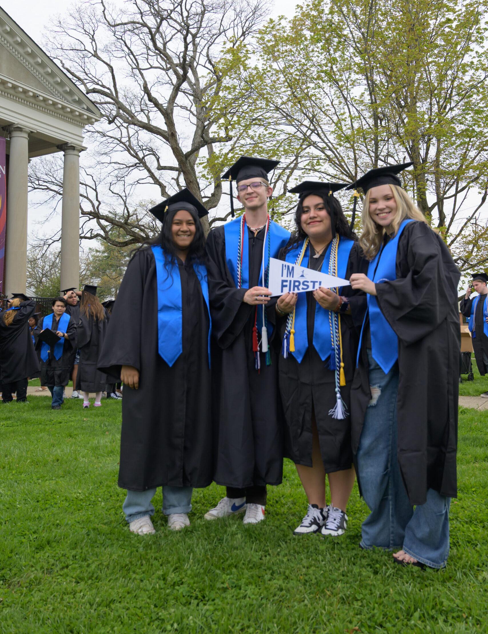 A group of four students who are standing outside on green grass, wearing black graduations caps and gowns, and blue stoles. The two students in the middle of the group are holding up a white pennant that reads 'I'm First' together.