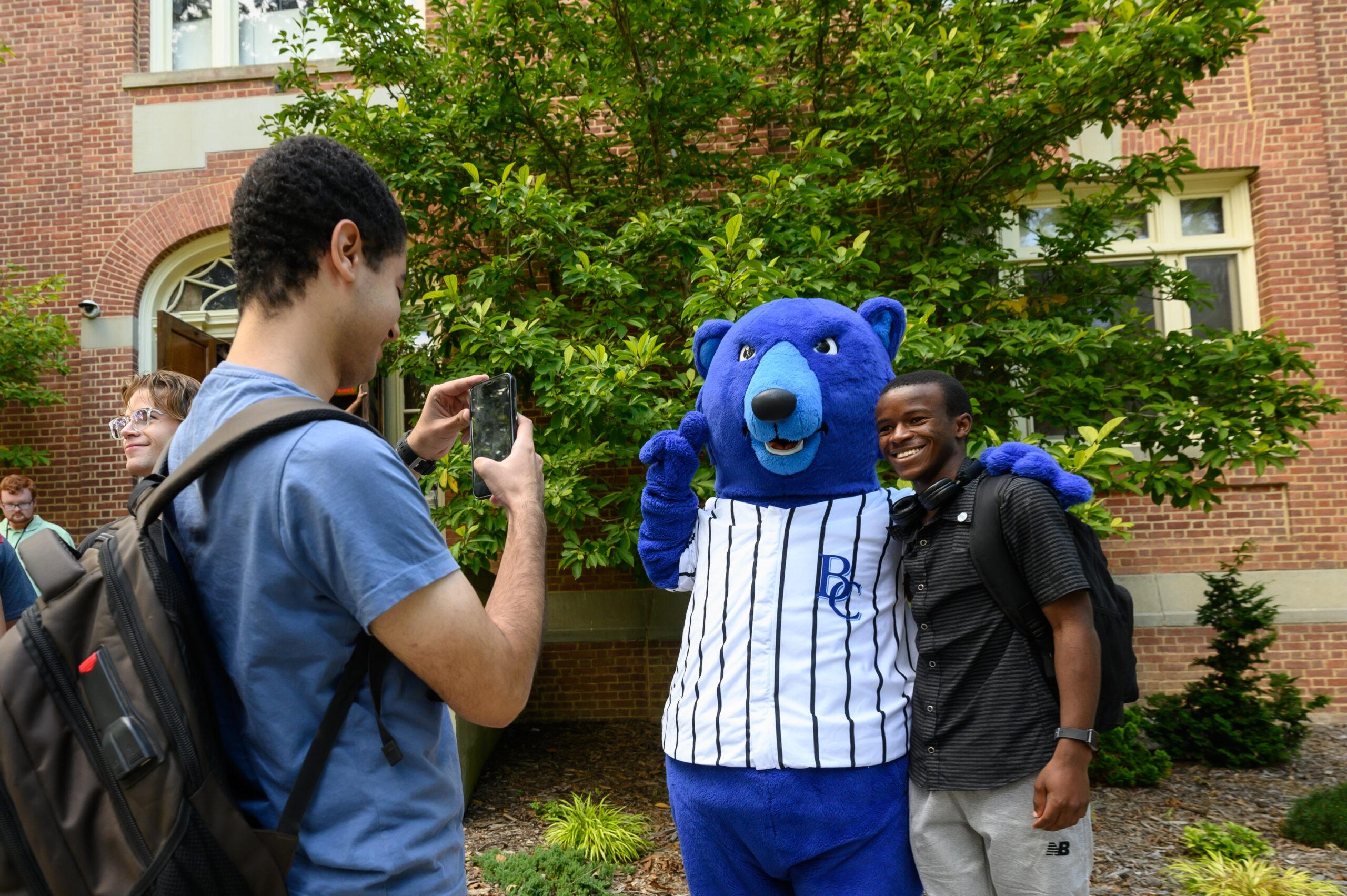 Student posing with a Blue the bear, the campus mascot while another student takes a photo outside a brick academic building.